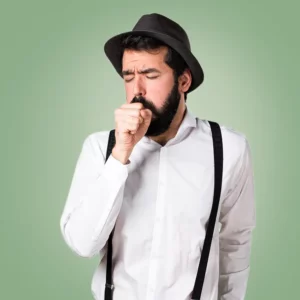 Dunkelhaariger Mann mit Bart, Hut und Hosenträgern über seinem weißen Hemd hustet in seine Faust