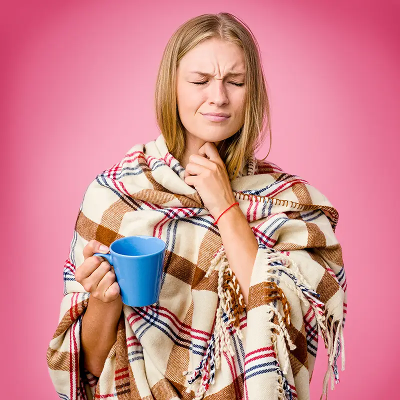 junge Frau hält sich ihre Hand an den Hals, ist in eine Decke eingewickelt und hat eine blaue Tasse in der Hand